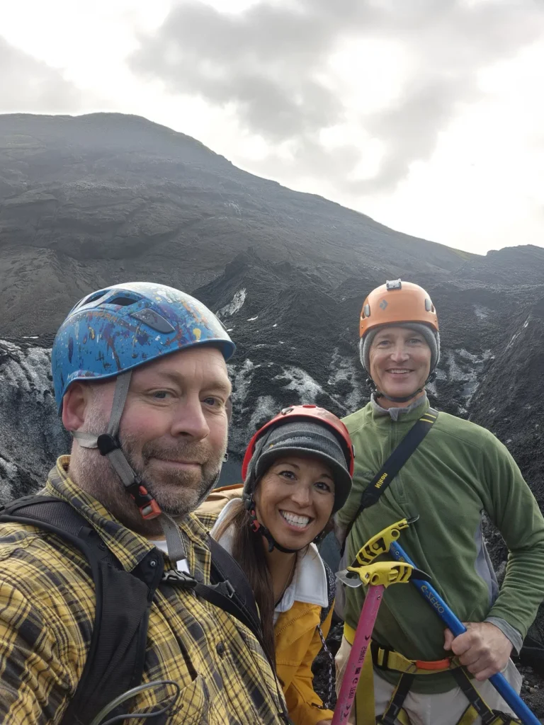 Three hikers in helmets pose for a selfie on a rocky Icelandic landscape.