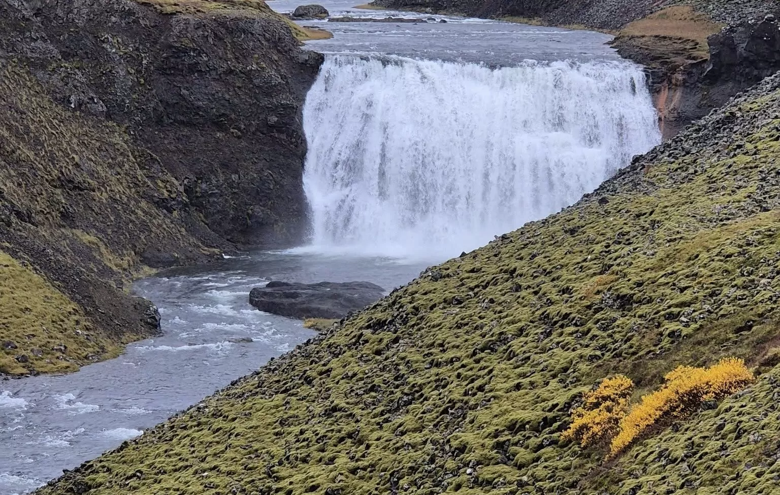Waterfall spilling into a river between moss-covered hills in Iceland.