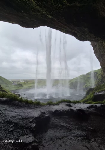 View from behind an Iceland waterfall looking out over green hills.