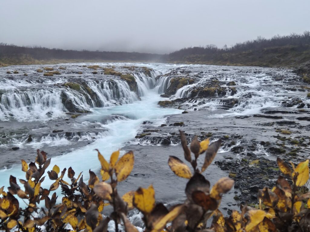 Wide view of Brúarfoss as bright blue glacial water cascades over rocky ledges under a misty sky.